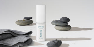 A bottle of Native dry shampoo powder, labeled "Cashmere & Rain," stands on a white surface next to stacked, wet stones and a gray towel, highlighting its non-aerosol, no butane dry shampoo formula