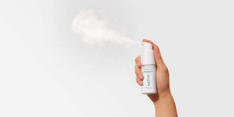 A hand holds a bottle of Native Dry Shampoo Powder, labeled "Butane Free, Talc Free," with a visible cloud of powder spraying from the nozzle, demonstrating how to use dry shampoo correctly for the best dry shampoo without residue or chemicals