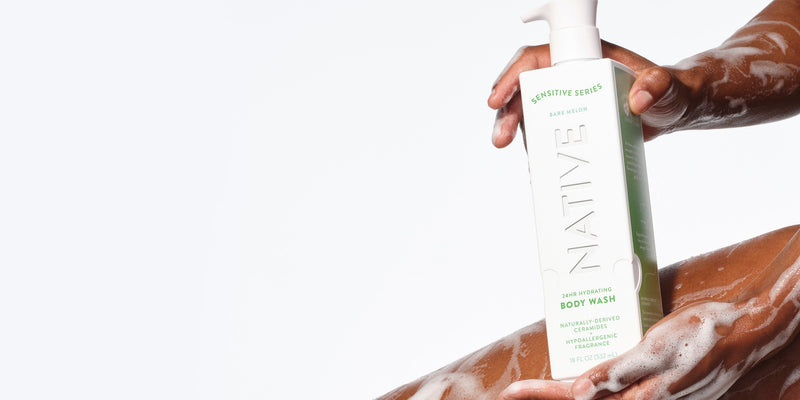 A person with dark skin holding a bottle of Native Sensitive Series Hydrating Body Wash, with visible suds on their arm, demonstrating how a moisturizing body wash for sensitive skin can help maintain the skin barrier and prevent dry skin