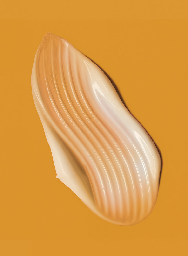 A glossy, textured smear of cream makeup, likely a liquid blush or cream bronzer, in a warm peach-beige shade, set against a solid mustard yellow background.