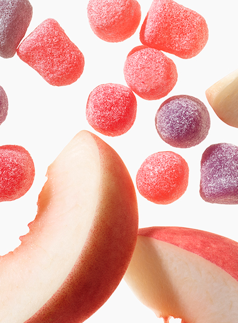 Sugar-coated peach-shaped candies in pink and purple floating above sliced peach pieces on a clean white background.