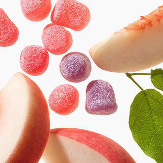 Sugar-coated peach-flavored gummy candies in pink and purple, artistically arranged with peach slices and a green leaf on a white background.