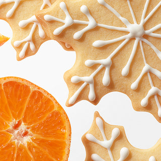 Close-up of decorated gingerbread cookies shaped like snowflakes and leaves with white icing, alongside a sliced orange on a white background.