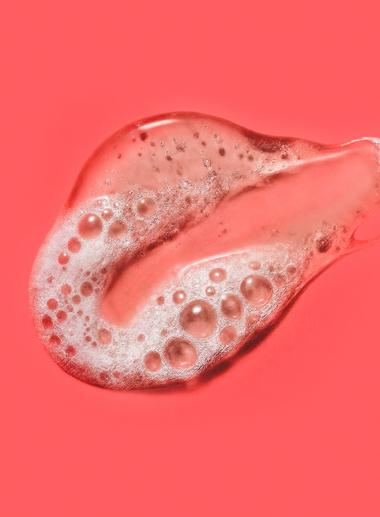 A close-up of a translucent pink cosmetic gel or serum swatch with white foam bubbles on a solid coral-pink background.