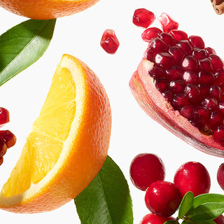 Fresh fruit arrangement on a white background, including a sliced orange with a green leaf, a pomegranate half showing red arils, loose pomegranate seeds, and red berries with green leaves.