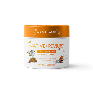 A 12 oz jar of Native x Peanuts Detoxifying Body Scrub in Maple Latte, featuring a white body with orange and black text and illustrations of Snoopy and Woodstock, and an orange lid.