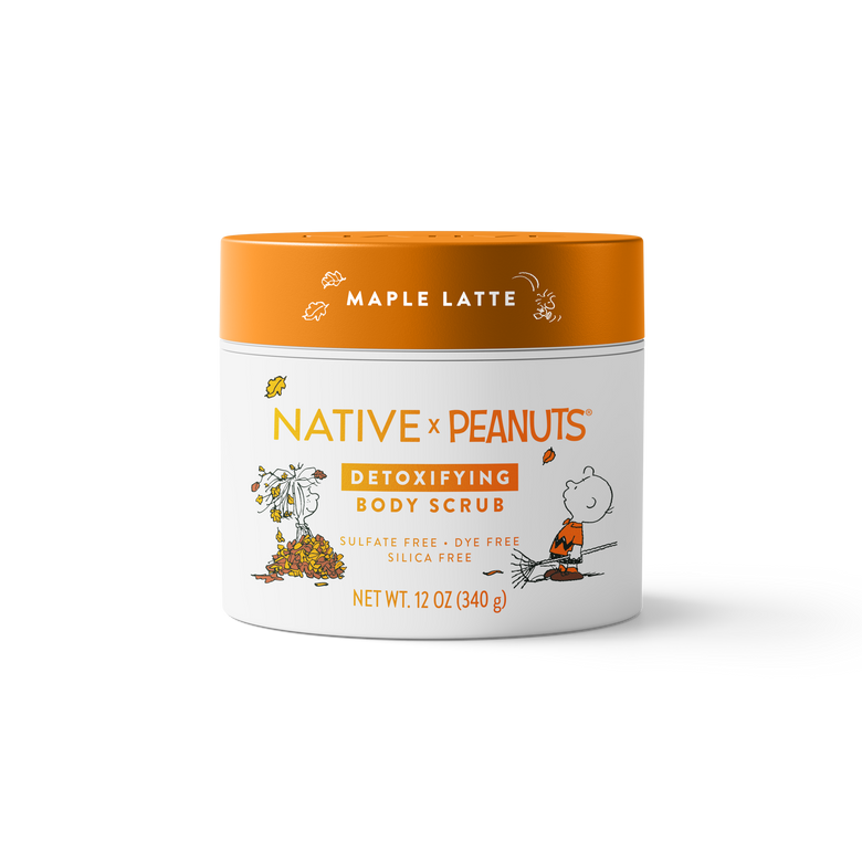 A 12 oz jar of Native x Peanuts Detoxifying Body Scrub in Maple Latte, featuring a white body with orange and black text and illustrations of Snoopy and Woodstock, and an orange lid.
