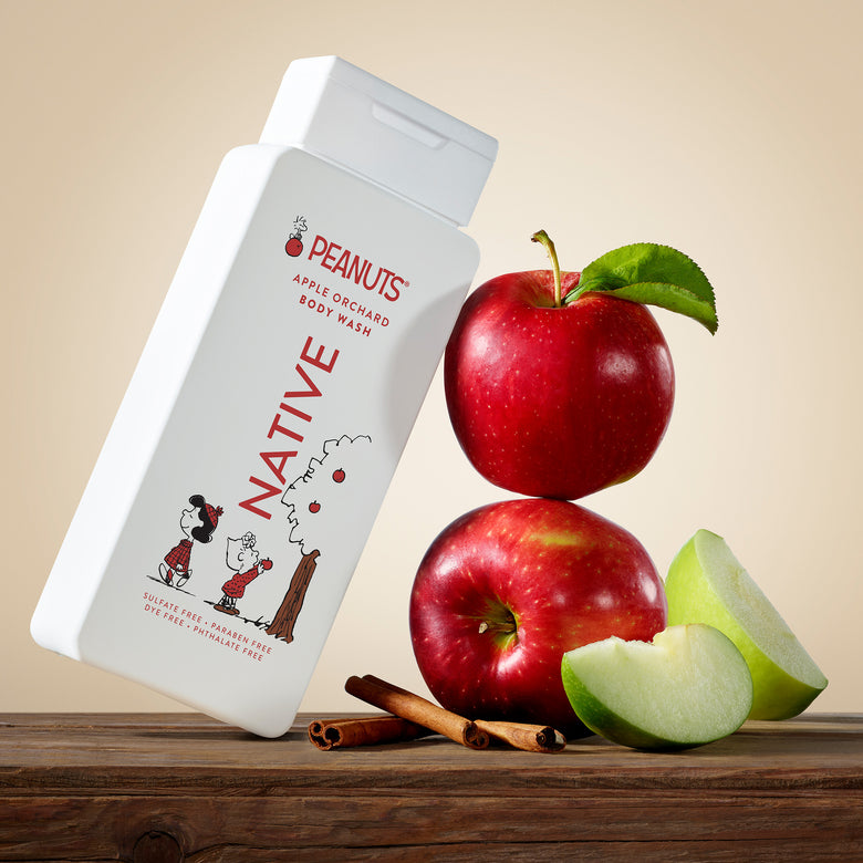 A bottle of Peanuts Native Apple Orchard Body Wash is displayed on a rustic wooden surface next to fresh red and green apples and cinnamon sticks, set against a warm beige background.