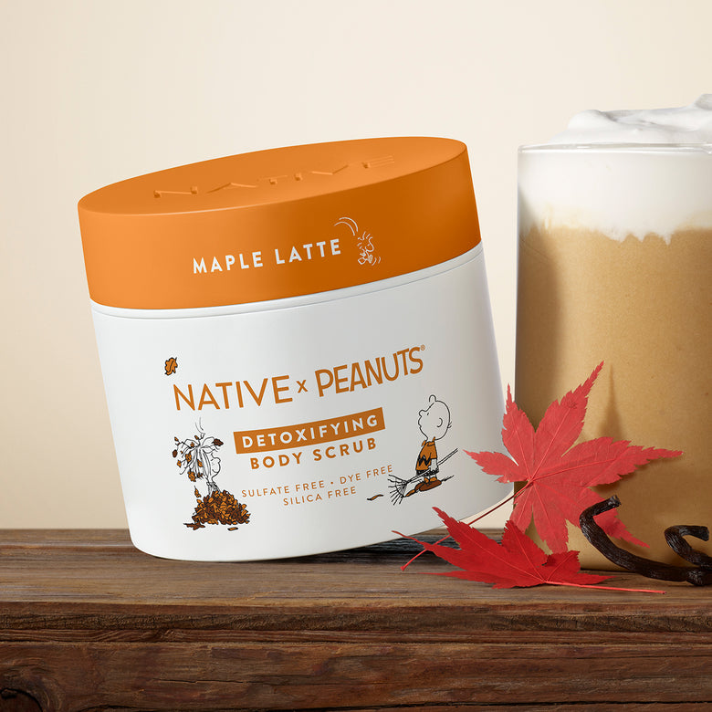 A tub of Native x Peanuts Detoxifying Body Scrub in Maple Latte scent, styled on a wooden surface next to a hot cocoa cup and red maple leaves.