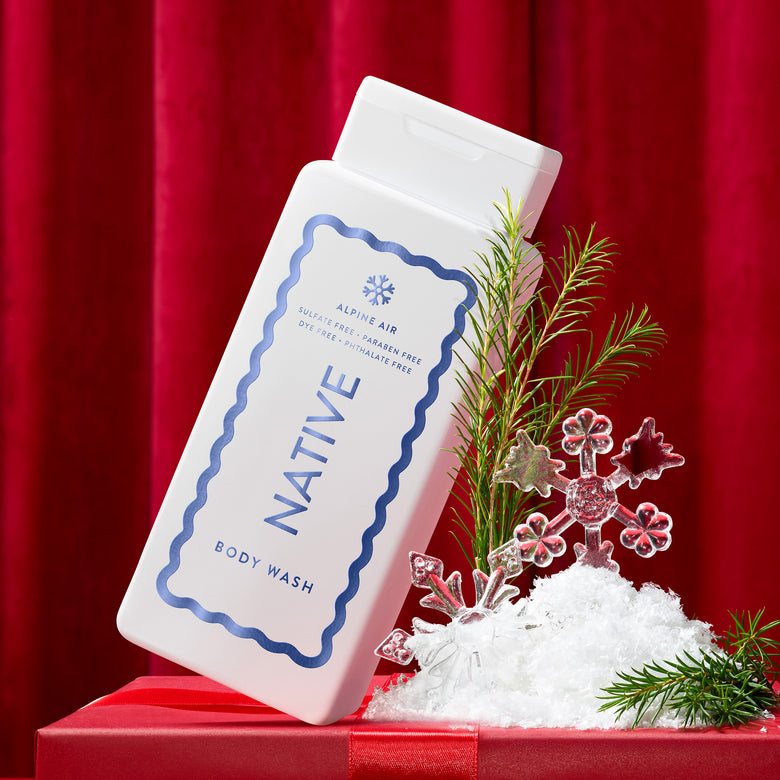 A white bottle of Native Alpine Air Body Wash, styled on a red gift box with white faux snow, green pine branches, and pink snowflake decorations against a red curtain background.