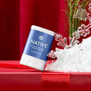 A Native brand aluminum-free deodorant stick in Alpine Air scent, styled on a red gift box with red ribbon next to white faux snow and holiday decorations against a red background.
