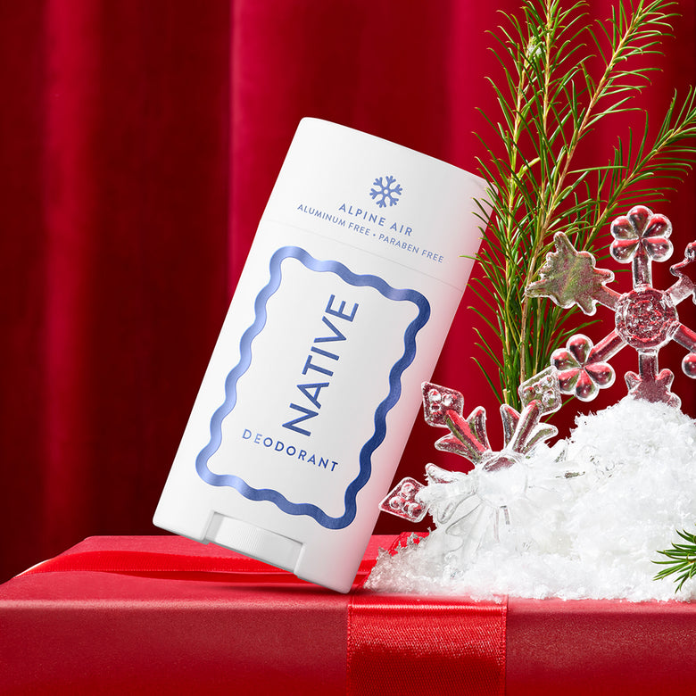 A white deodorant stick labeled NATIVE DEODORANT with blue text and border, placed on a red gift box with red fabric backdrop, white faux snow, and green pine branches. The product itself is the sold item; surrounding props are decorative.