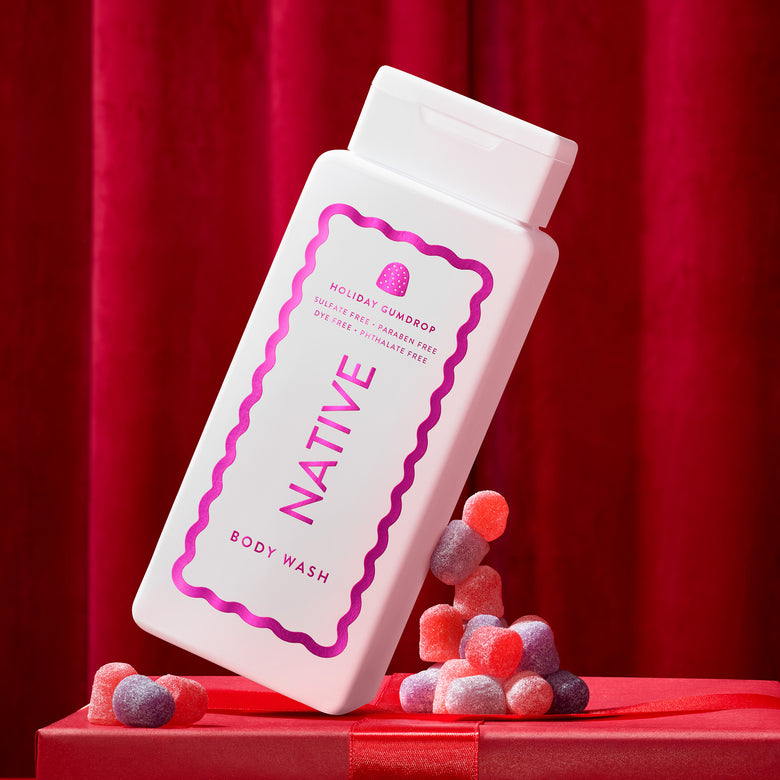 A white bottle of Native Body Wash with a pink wavy border and strawberry graphic, shown on a red gift box with candy against a red curtain background.