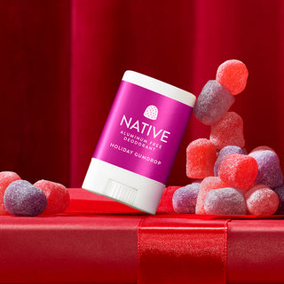 A stick of Native Holiday Gumdrop aluminum-free deodorant is shown tilted on top of a red gift box with a red ribbon, surrounded by colorful sugar-coated gumdrops against a red draped background.