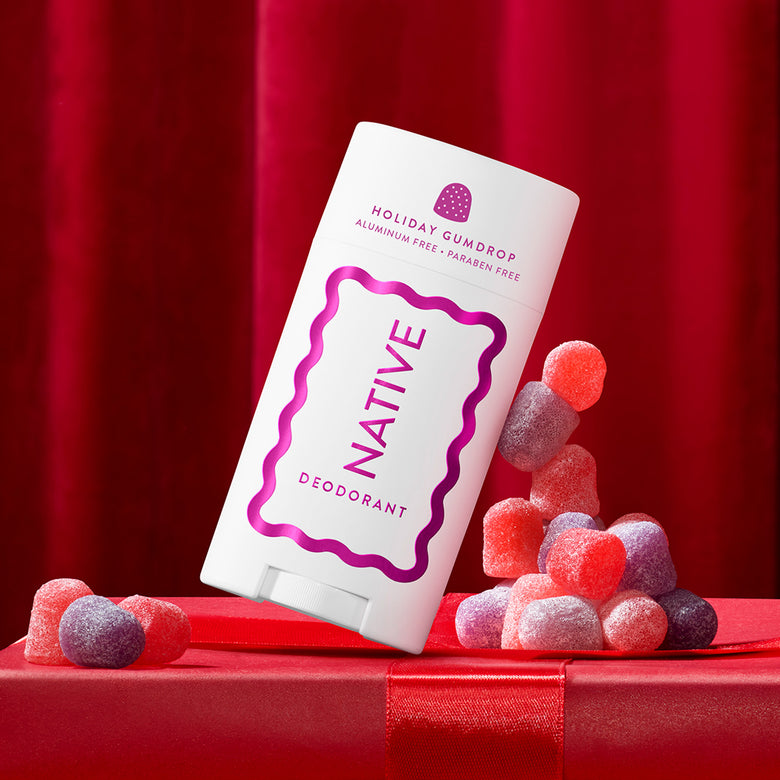 A white Native Deodorant stick package with purple text and border graphics, styled on a red gift box with red and purple gumdrops against a red curtain background.