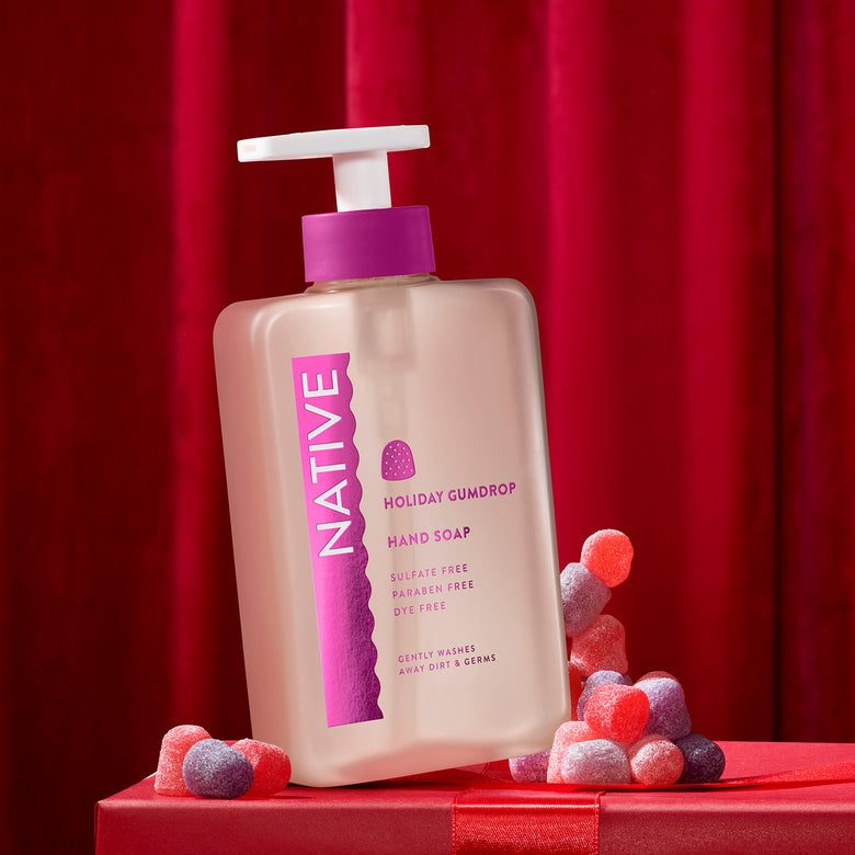 A bottle of Native Holiday Gumdrop hand soap stands on a red gift box against a red curtain backdrop, with red and pink candy pieces scattered around the base.