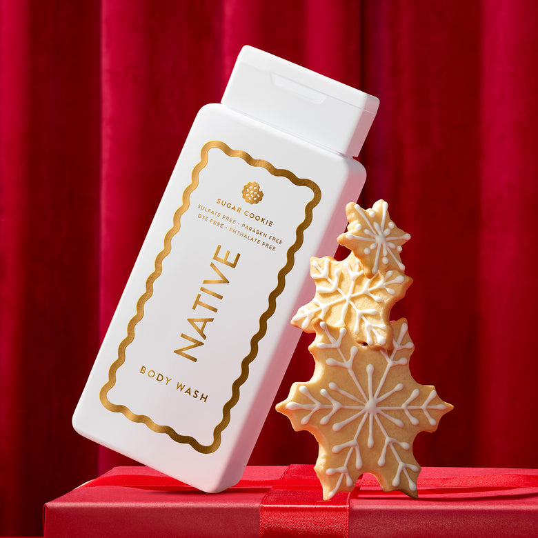 A white bottle of Native Sugar Cookie body wash with gold lettering and border details, styled on a red gift box against a red curtain backdrop next to three decorated gingerbread cookies shaped like snowflakes and a tree.