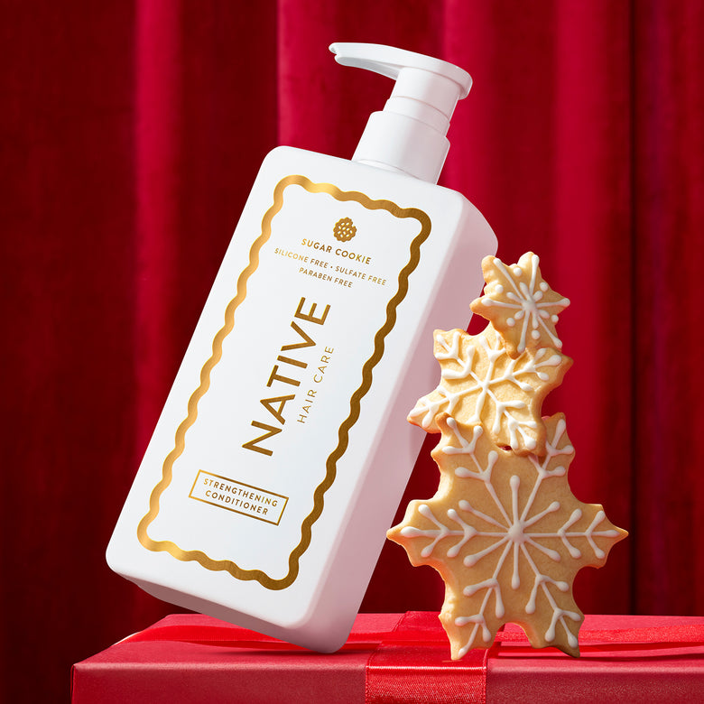 A white pump bottle of Native Hair Care Sugar Cookie Strengthening Conditioner with gold lettering, styled next to decorated sugar cookie snowflakes on a red gift box against a red curtain background.