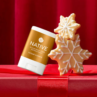 A Native brand deodorant stick labeled 'Sugar Cookie' stands on a red gift box next to two decorated sugar cookie snowflakes against a red draped background.