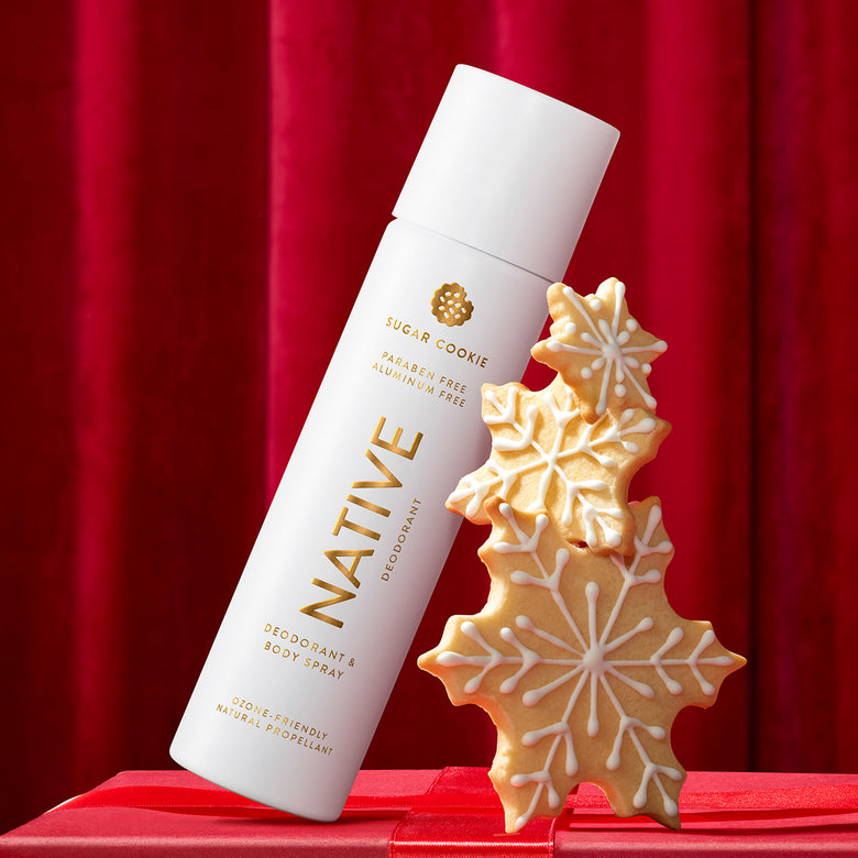 A white can of Native Sugar Cookie deodorant and body spray with gold lettering, styled next to three decorated sugar cookie snowflakes on a red gift box against a red curtain backdrop.