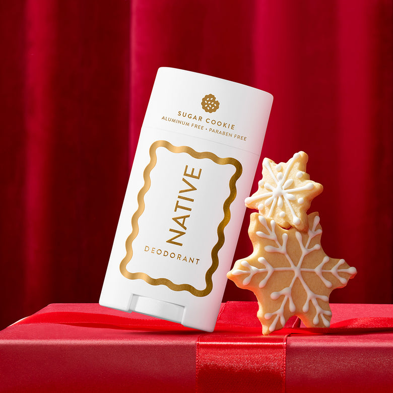 A white Native Deodorant stick labeled 'Sugar Cookie' stands on a red gift box with two decorated sugar cookie snowflake shapes beside it against a red curtain backdrop.