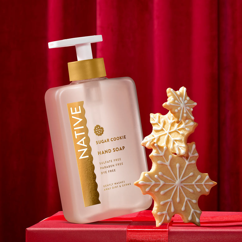 A bottle of Native Sugar Cookie hand soap stands on a red gift box next to three decorated gingerbread cookie snowflake shapes against a red curtain background.