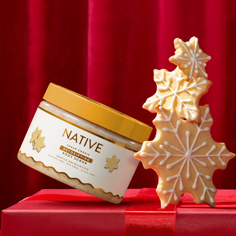 A jar of Native Sugar Cookie Gifted Skin Body Scrub with a gold lid and white label, styled next to decorated snowflake cookies on a red gift box against a red curtain background.