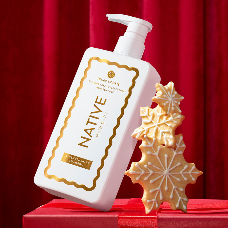 A white pump bottle of Native Hair Care Sugar Cookie shampoo with gold lettering, styled on a red gift box next to three decorated sugar cookie snowflakes against a red curtain backdrop.
