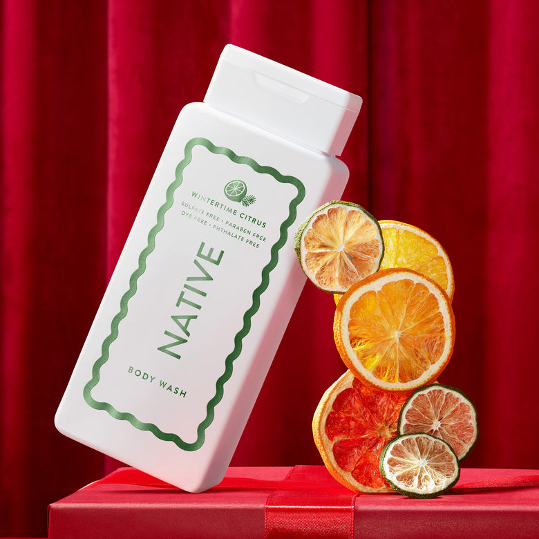 A white bottle of Native body wash with a green label, styled on a red gift box with dried citrus slices against a red curtain background.