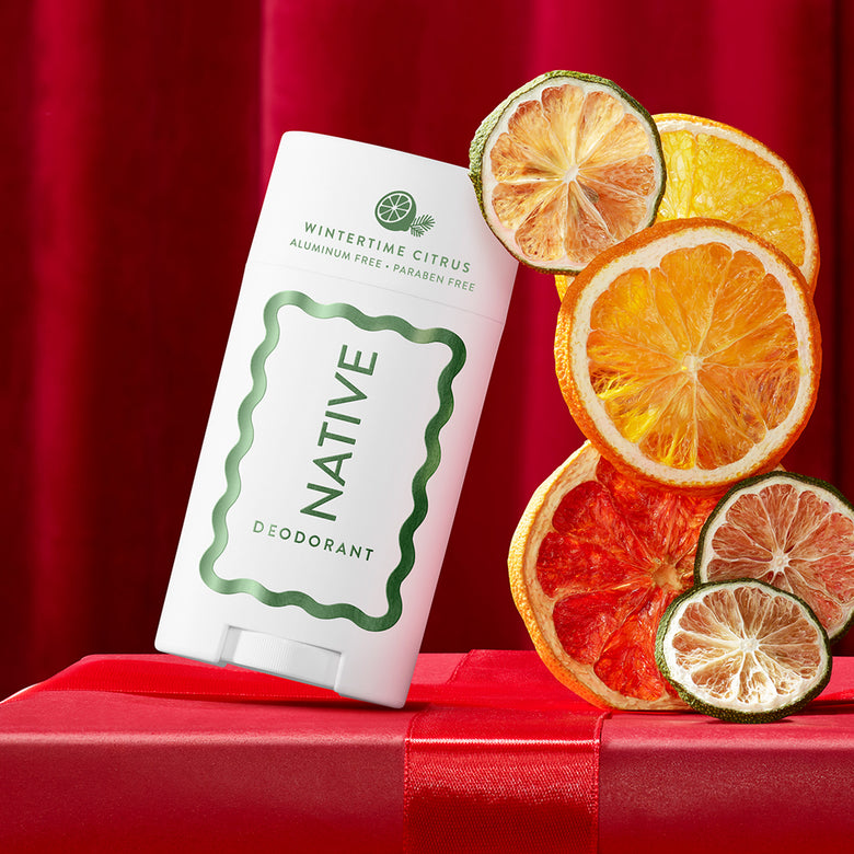 A Native Deodorant stick in Wintertime Citrus scent, displayed on a red gift box with dried citrus slices including orange, grapefruit, and lime against a red curtain background.