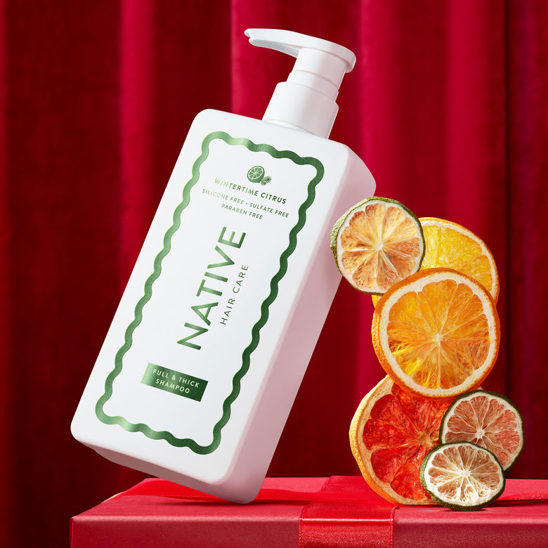 A bottle of Native Hair Care shampoo with a white pump and green label, styled on a red box against a red curtain backdrop, with dried orange and lime slices beside it.