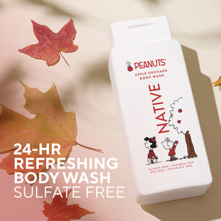 A white bottle of Peanuts Apple Orchard Body Wash by Native, styled with autumn leaves on a warm beige background. The packaging features red and black text and character illustrations of Snoopy and Charlie Brown.