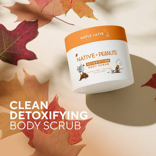 A jar of Native x Peanut Detoxifying Body Scrub labeled Maple Latte, styled with autumn maple leaves on a warm beige background. Large text on the left reads 'CLEAN DETOXIFYING BODY SCRUB'.