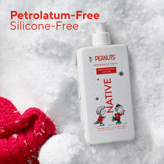 A white bottle of Native Peanuts Peppermint Patty moisturizing lotion sits on a white textured surface with a red knit heart-shaped accessory at the bottom left. Red text at the top reads 'Petrolatum-Free Silicone-Free'.