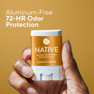 A hand holding a Native brand aluminum-free deodorant stick in the Sugar Cookie scent, set against a warm golden-brown background with white text reading 'Aluminum-Free 72-HR Odor Protection'.