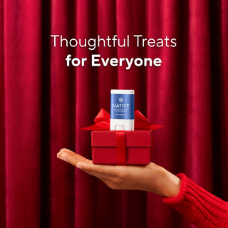 A hand in a red knit sweater holds a small red gift box with a red ribbon and a Native brand product stick on top, set against a red curtain backdrop with the text 'Thoughtful Treats for Everyone'.