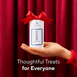 A hand holds a white bottle of Alpine Air Native D-Isboquant deodorant, tied with a red ribbon, against a red curtain background with the text 'Thoughtful Treats for Everyone'.