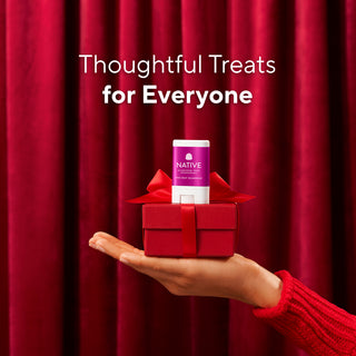A hand in a red knit sweater holds a small red gift box with a red ribbon and a Native brand product stick on top, set against a red curtain backdrop with the text 'Thoughtful Treats for Everyone'.