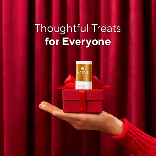 A hand in a red knit sweater holds a small red gift box with a red ribbon and a Native brand product stick centered on top, against a red curtain backdrop with the text 'Thoughtful Treats for Everyone'.