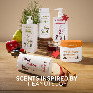 A collection of Native Peanuts body care products arranged on a wooden tabletop with seasonal props including apples, berries, cinnamon sticks, pine branches, and a small glass jar. Visible products include pump bottles, a tub, and a roll, all branded Native Peanuts, with one tub labeled Maple Latte.