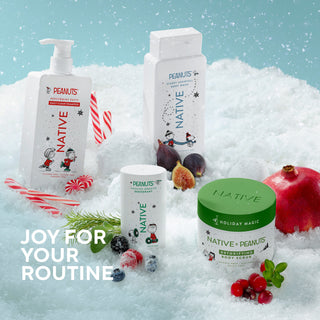 Four Native Peanuts holiday body care products arranged in white faux snow with candy canes, figs, blueberries, pomegranate, and greenery against a light blue snowy background. Visible products include a pump bottle, a tall box, a small tub, and a jar labeled Native Peanuts, with the text 'JOY FOR YOUR ROUTINE' on the left.