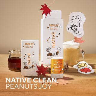 A set of Native Clean body products themed around Peanuts, including a maple latte body wash, deodorant stick, and mini deodorant stick, styled on a wooden table with autumn leaves, a mug of coffee, and cartoon character stickers.