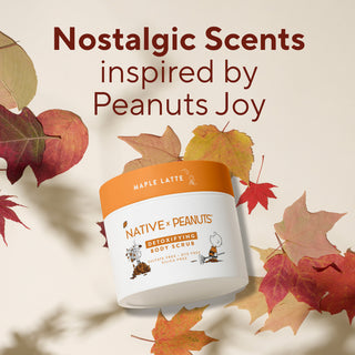 A jar of Native Peanuts Detoxifying Body Scrub labeled Maple Latte, styled with autumn leaves on a warm beige background with promotional text reading 'Nostalgic Scents inspired by Peanuts Joy.'.